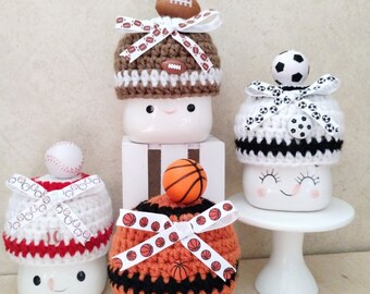 May include: Four white ceramic mugs with different sports-themed crocheted hats. The hats are decorated with a football, soccer ball, baseball, and basketball. The mugs have a happy face.