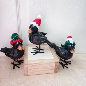 May include: Three black bird figurines wearing red and green crocheted Santa hats. The birds are standing on a wooden base.