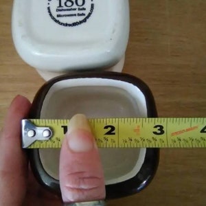 May include: A brown and white ceramic square dish with a black rim. The dish is 3 inches wide. The bottom of the dish has a white sticker with the text "One Hundred 80 degrees.com" and "180 Dishwasher Safe Microwave Safe".