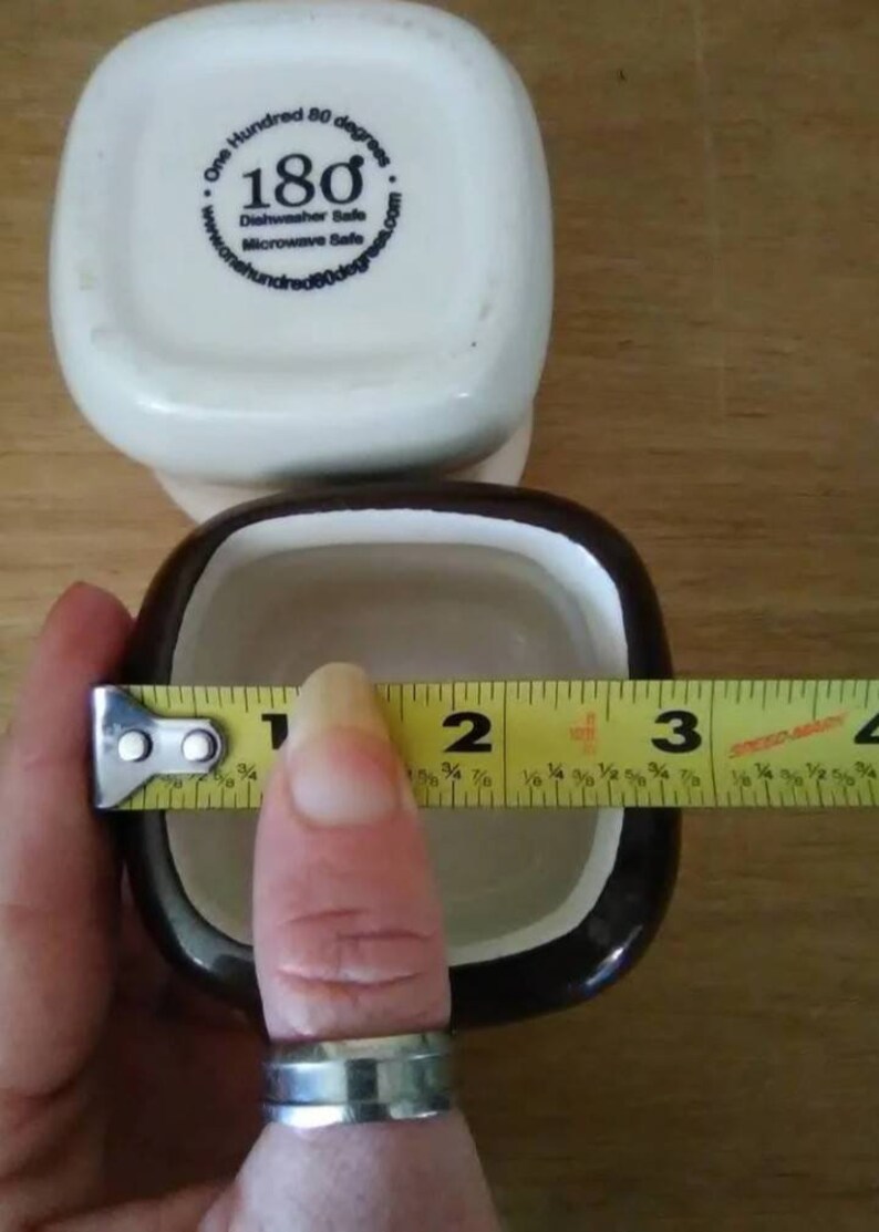 May include: A brown and white ceramic square dish with a black rim. The dish is 3 inches wide. The bottom of the dish has a white sticker with the text "One Hundred 80 degrees.com" and "180 Dishwasher Safe Microwave Safe".