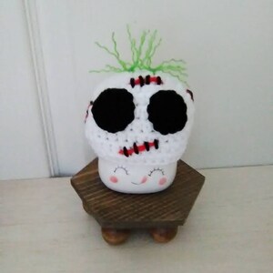May include: White ceramic mug with a smiling face and black eyes. The mug is wearing a white crocheted hat with black eyes, red stitching, and green yarn hair.