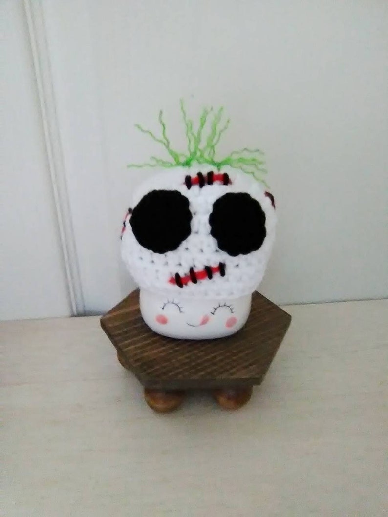 May include: White ceramic mug with a smiling face and black eyes. The mug is wearing a white crocheted hat with black eyes, red stitching, and green yarn hair.