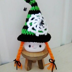 May include: A white ceramic mug with a cute face, wearing a black and green striped witch hat with a white crocheted spiderweb and black spiders. The mug has orange yarn braids tied with black ribbon.