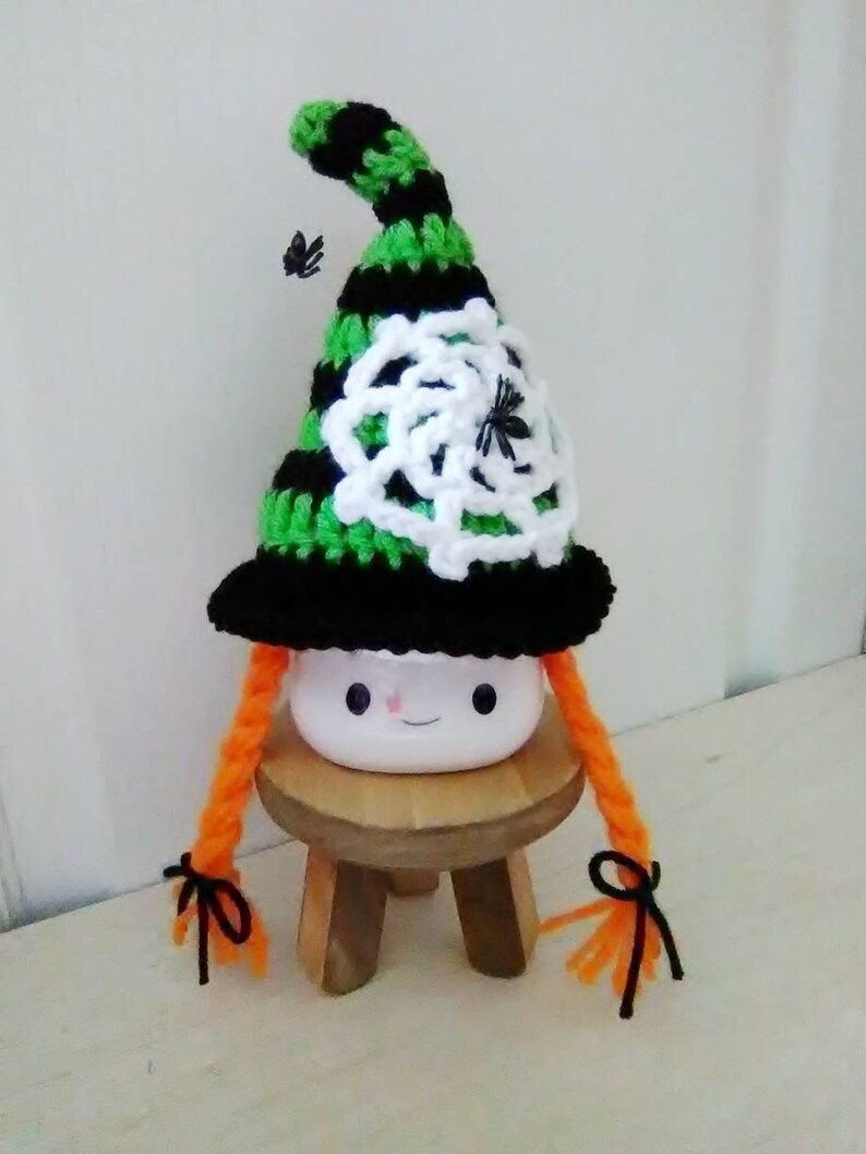 May include: A white ceramic mug with a cute face, wearing a black and green striped witch hat with a white crocheted spiderweb and black spiders. The mug has orange yarn braids tied with black ribbon.