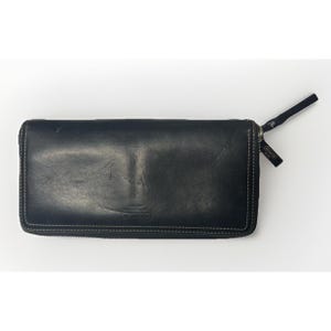 May include: Black leather wallet with white stitching and a zipper closure. The wallet has a wrist strap and a small tag with the brand name. The wallet is rectangular and appears to be made of a smooth leather material.