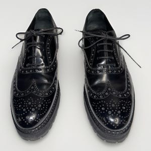 May include: Pair of black leather Oxford shoes with a glossy finish. The shoes feature decorative perforations and a thick, lug sole. The shoes have black laces and a classic wingtip design.