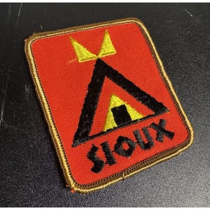 May include: A rectangular embroidered patch with a bright red background and a gold-colored border. The design features a black triangular tent shape with a yellow entrance and two yellow triangular accents above it, with the word SIOUX in black.
