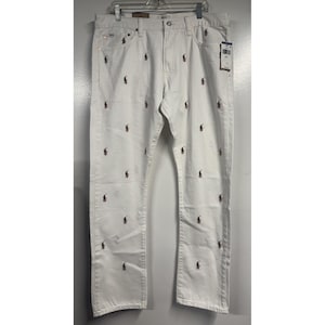May include: Cream-coloured denim trousers featuring a repeating pattern of small red polo player logos. The trousers have a classic five-pocket design and a straight leg fit. A tag is attached.