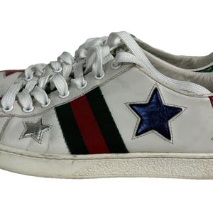 May include: A white low-top sneaker featuring a central black and red vertical stripe, white laces, and metallic star appliqués in red, silver, blue, and gold. The heel tab is finished in a textured red metallic material with a green accent.