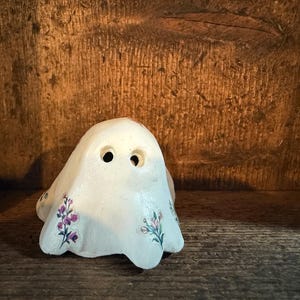 May include: A small, white ghost figurine with two eye holes and floral accents in purple and green. The ghost is sitting on a wooden surface, with a wooden background.