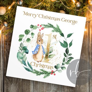 May include: A white card with a gold glitter background and a watercolor illustration of a rabbit wearing a blue jacket. The card says "Merry Christmas George 1st Christmas".