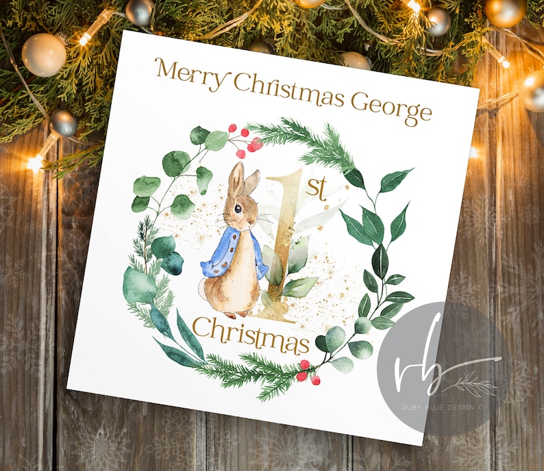 May include: A white card with a gold glitter background and a watercolor illustration of a rabbit wearing a blue jacket. The card says "Merry Christmas George 1st Christmas".
