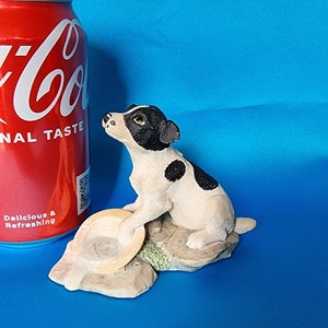 May include: A white and black figurine of a Jack Russell terrier dog sitting on a rock with a brown dish in front of it.