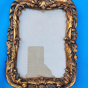 May include: A gold-colored ornate picture frame with a decorative design of leaves and swirls. The frame has a glass front and a beige mat.