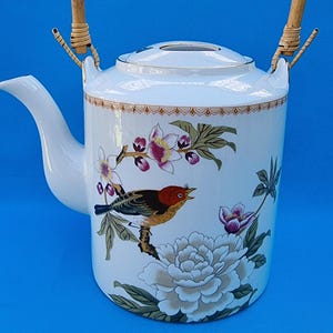 May include: White porcelain teapot with a floral design featuring a bird, a large white flower, and pink flowers. The teapot has a bamboo handle.