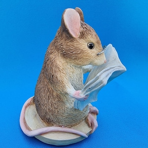 May include: A detailed figurine of a mouse reading a paper, wearing small spectacles. The mouse is brown and tan with pink ears and feet. It sits on a light-coloured base against a bright blue background. The mouse appears to be engrossed in the paper.