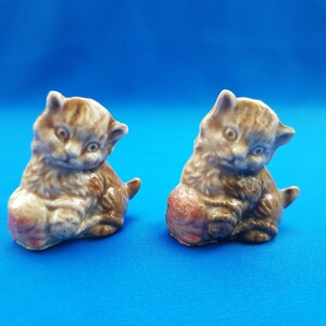 Puede incluir: Dos figuritas de gatitos de cerámica marrón y blanco, cada una sosteniendo una bola de lana. Los gatitos están sentados sobre una superficie azul.