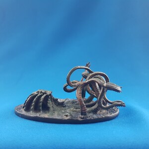 May include: A miniature painted figure of a tentacled creature with a dark green and brown color scheme. The creature is posed on a circular base with a rocky texture.