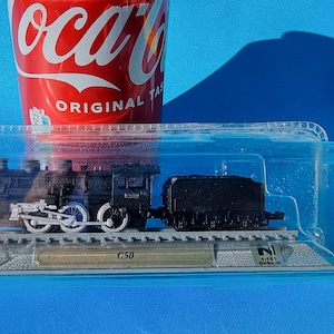 May include: A black model train engine and tender in a clear plastic display case with a gold label that reads "C50". The train is on a track and there is a small Japanese flag on the display case.