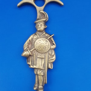 Könnte beinhalten: Ein Kaminbesteck aus Messing mit einer dekorativen Figur eines Soldaten, der einen Schild und einen Speer hält. Der Kaminbesteck hat oben einen Haken und einen langen, dünnen Schaft.