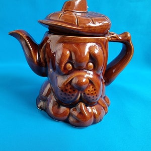 Könnte beinhalten: Eine braune Teekanne aus Keramik in Form eines Hundes, der einen braunen Hut trägt. Die Teekanne hat einen Ausguss und einen Griff.
