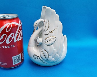 May include: A white ceramic swan-shaped vase with gold accents, including leaves and a flower, stands next to a red Coca-Cola can. The vase has a detailed design and is set against a blue background.