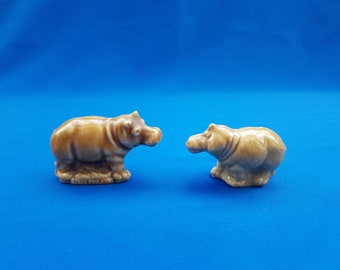 May include: Two brown ceramic hippopotamus figurines. The hippopotamus on the left is standing with its legs spread apart. The hippopotamus on the right is sitting with its legs tucked under its body.