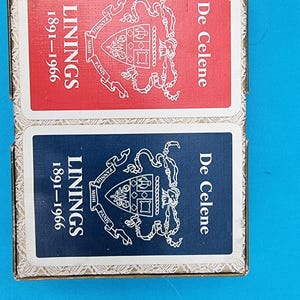 Peut inclure: Une boîte de cartes à jouer vintage avec un design rouge et bleu. La boîte porte l'inscription "Linings 1891-1966" et "De Celene" sur le devant.