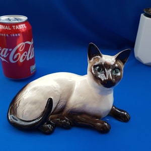 Op de afbeelding: Een keramiek beeldje van een liggende Siamese kat. De kat is wit met bruine markeringen op zijn gezicht, oren en staart. De kat heeft groene ogen en een zwarte neus.