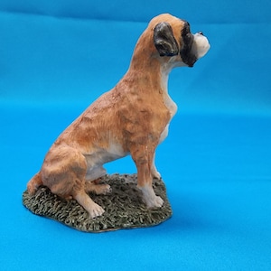 Puede incluir: Una figurita de cerámica marrón y blanca de un perro boxer sentado sobre una base verde. El perro mira hacia la derecha.