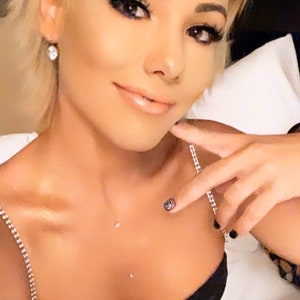 May include: A woman with short blonde hair wearing a black lace top with a silver chain strap. She is wearing a diamond earring and has a small diamond piercing on her chest. Her nails are painted black with a small design on one finger.