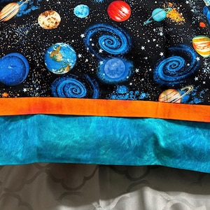 Op de afbeelding: Een stoffen item met een ruimtethema. Het bovenste gedeelte toont planeten, sterrenstelsels en sterren tegen een zwarte achtergrond. Een oranje streep scheidt het ruimteontwerp van een turquoise onderkant.