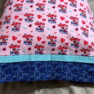 Könnte beinhalten: Ein rosa Kissenbezug mit einem sich wiederholenden Muster aus rotem und blauem Text mit der Aufschrift "Suck It You", Herzen und kleinen blauen Kreisen. Der untere Rand hat ein blaues Herzmuster.