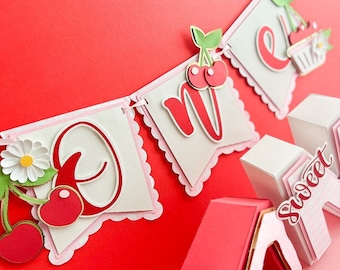 May include: A white and pink banner with the word "One" and a cherry design. The banner is decorated with red and green cherries. The banner is attached to a white and pink box with the word "Sweet" on it.