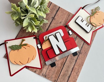 May include: A red truck with a white "N" on the side, carrying a small orange pumpkin. The truck is part of a banner with a white background and red trim. The banner also features a large orange pumpkin with a green stem and leaves. The banner says "ONE" in white letters.