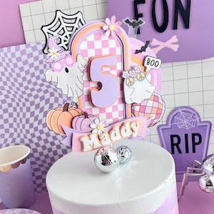 May include: A layered cake topper featuring a large number 5, the name Maddy, ghosts, pumpkins, a disco ball, and a spider web in pastel pink, purple, and orange. The topper is displayed on a white, purple, and pink ombre cake with gold flecks.