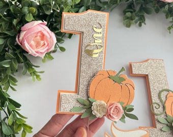 May include: A cake topper featuring a glittery gold number one with the word "one" written in gold script. The number one is decorated with a small orange pumpkin, green leaves, and pink flowers. The topper also includes a white banner with the word "party" written in gold script.