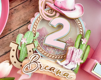 May include: Pink and green cake topper with a number 2, a cowboy hat, a horseshoe, cacti, and the name Breanna. The topper is decorated with glitter and a rope design.