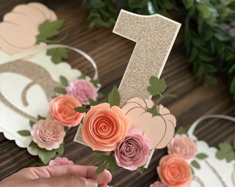 May include: A white banner with a gold glitter number one, a pink and orange paper flower arrangement, and a pink pumpkin. The banner is decorated with green leaves and has a scalloped edge.