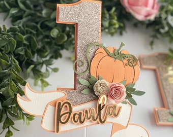 May include: A glittery gold number one cake topper with a pumpkin and flowers. The topper has a white banner with the name "Paula" written in gold.