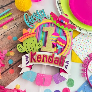 May include: A colorful fiesta themed cake topper with a cactus, sombrero, and the number one. The topper also includes the name Kendall and a banner with the word "fiesta".