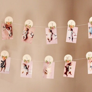 May include: A string of white scalloped circles with a floral design and a silver glitter number on each circle. Each circle is attached to a string with a clothespin and a photo of a baby. The photos are arranged in order from 1 to 11. The photos are all on a pink background.