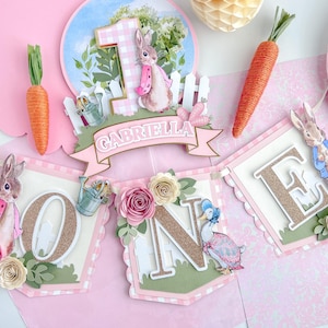 May include: A pink and green banner with the word "ONE" in gold glitter letters. The banner features a Peter Rabbit character, a duck, and flowers. The banner also includes the name "Gabriella".