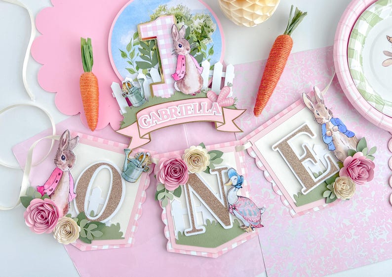 May include: A pink and green banner with the word "ONE" in gold glitter letters. The banner features a Peter Rabbit character, a duck, and flowers. The banner also includes the name "Gabriella".
