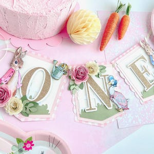 May include: A pink and green banner with the word "ONE" in gold glitter letters. The banner is decorated with flowers, a rabbit, a duck, and a watering can. The banner is perfect for a first birthday party.