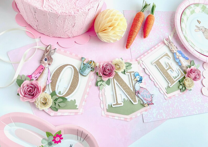 May include: A pink and green banner with the word "ONE" in gold glitter letters. The banner is decorated with flowers, a rabbit, a duck, and a watering can. The banner is perfect for a first birthday party.