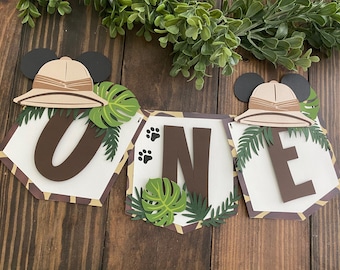 Può includere: Uno striscione di carta marrone e bianco con la parola "ONE" in lettere marroni. Lo striscione è decorato con foglie verdi, impronte di zampe marroni e due orecchie marroni di Topolino con un cappello da safari beige.