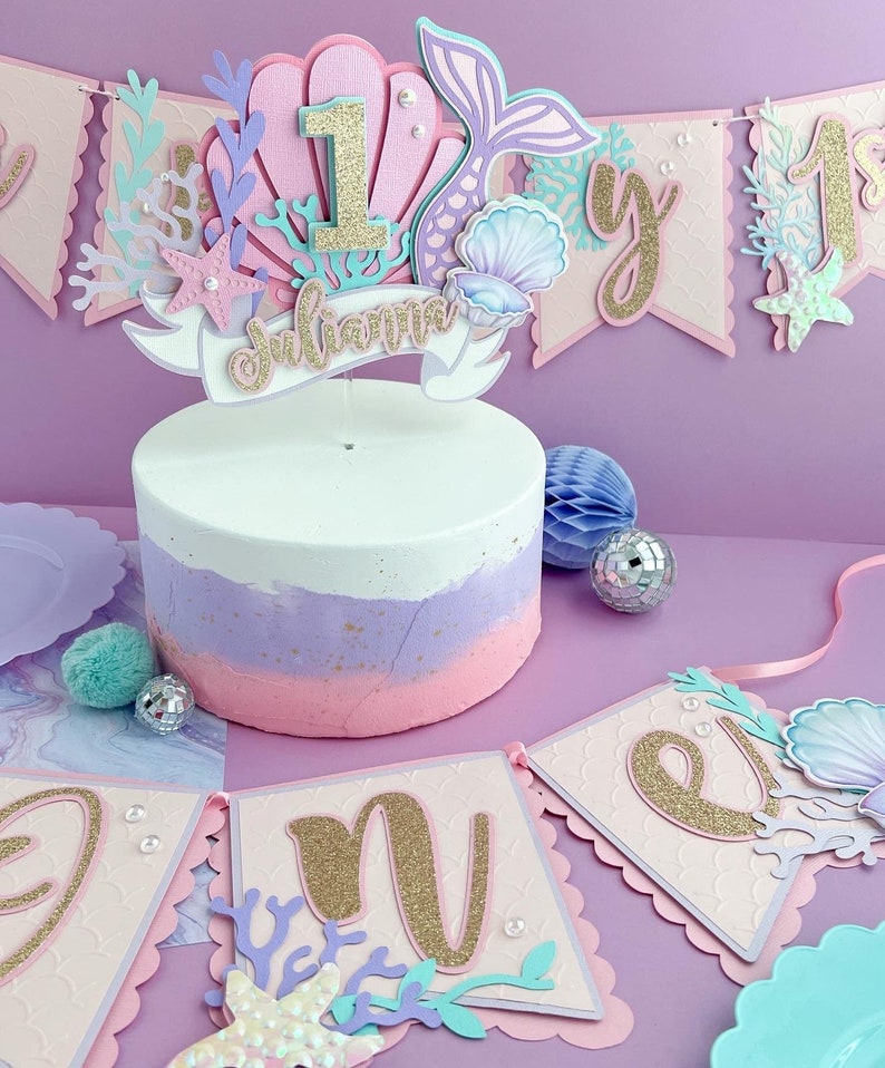 Peut inclure: Un topper de g&acirc;teau sur le th&egrave;me de la sir&egrave;ne rose et violet avec un num&eacute;ro un en paillettes dor&eacute;es, une queue de sir&egrave;ne, des coquillages et une banni&egrave;re qui dit "Julianna".