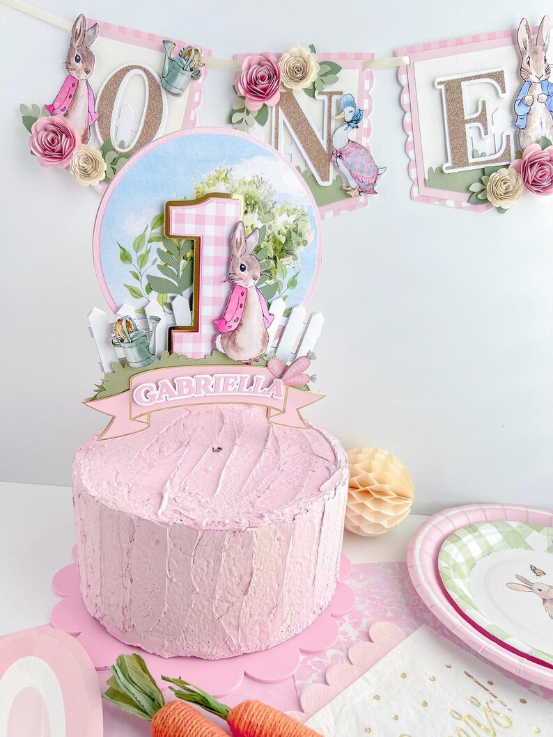 May include: A pink and white cake with a bunny and the number one on top. The cake is decorated with a banner that says "ONE" and has a pink and white gingham pattern. The cake is also decorated with a banner that says "Gabriella".