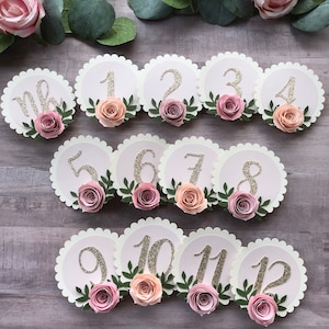 May include: Twelve white scalloped circles with pink and peach paper flowers and green leaves. Each circle has a gold glitter number from one to twelve.